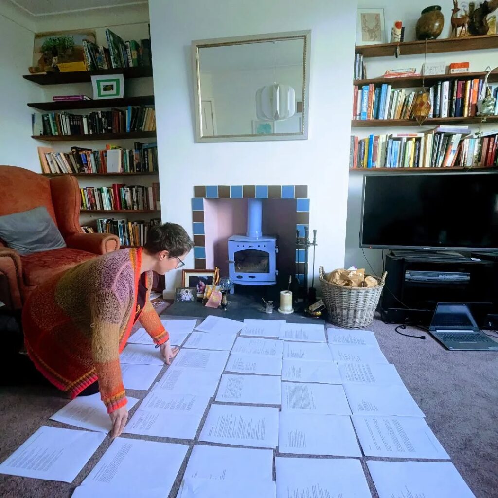 Becky kneeling, looking at poems laid out on the carpet. A wood burner and book shelves are in the background. 