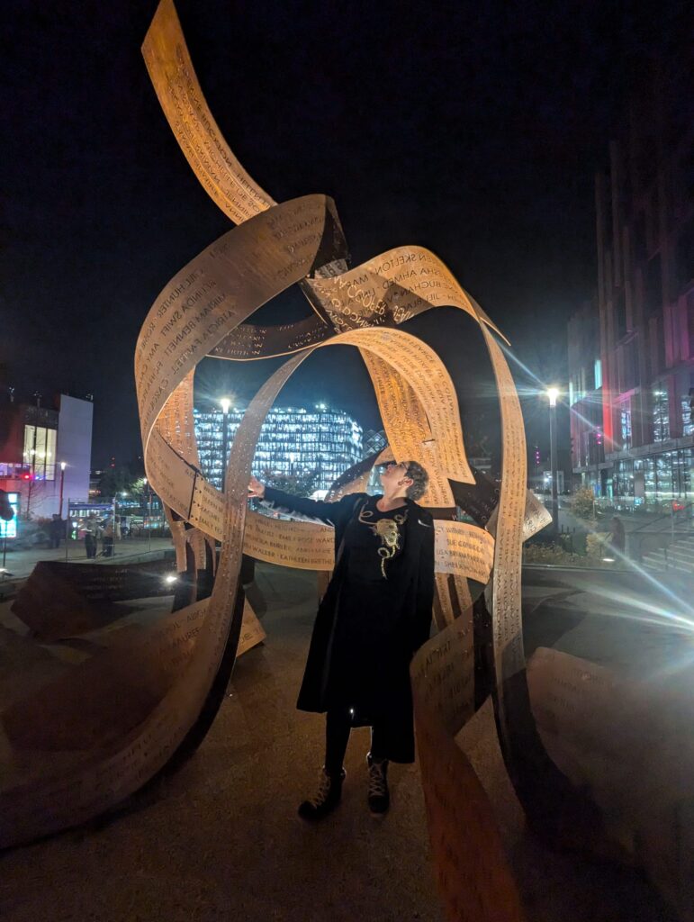 Becky looks up, touching Pippa Hale's Ribbons Sculpture. The work is lit with Leeds buildings in the background including The Leeds Playhouse. Photograph by Abdelrahman Hegazy.