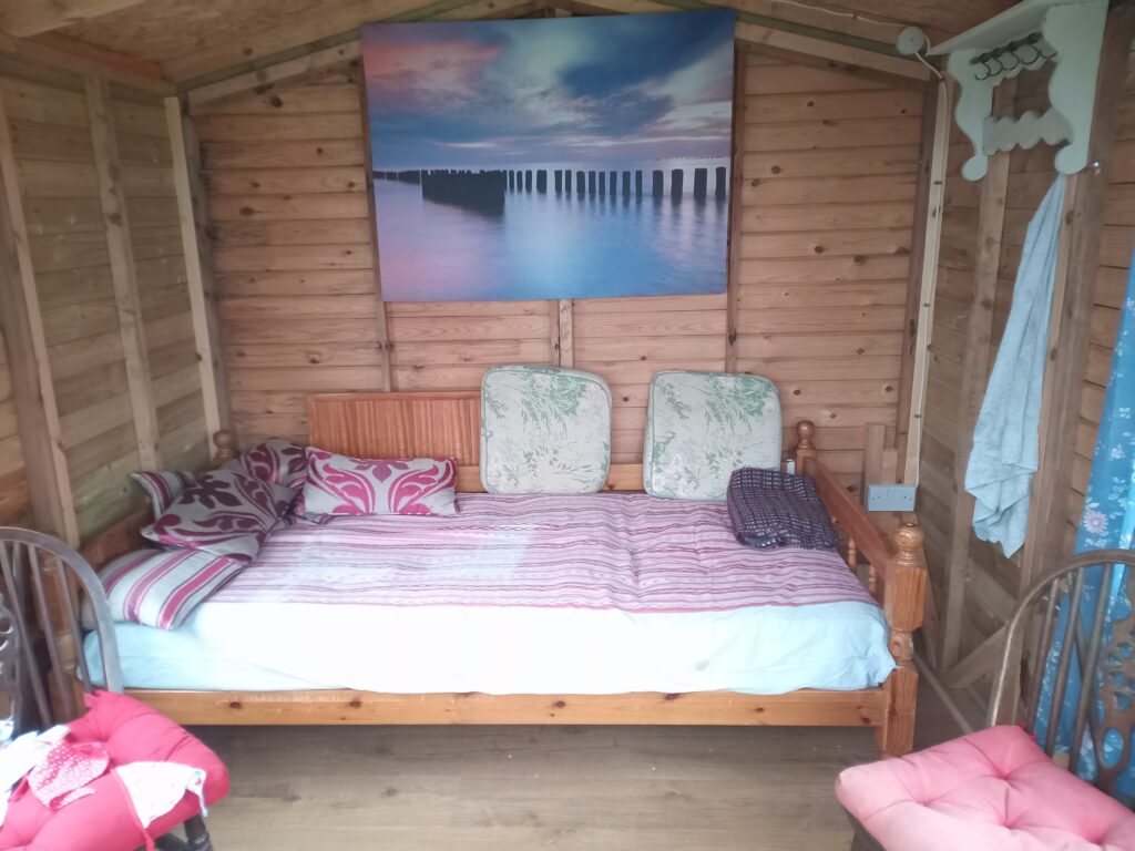 Inside a wooden hut with a double bed with cushions and throw. A canvas of groynes in the sea hangs on the wall.  Two chairs are in the foreground.