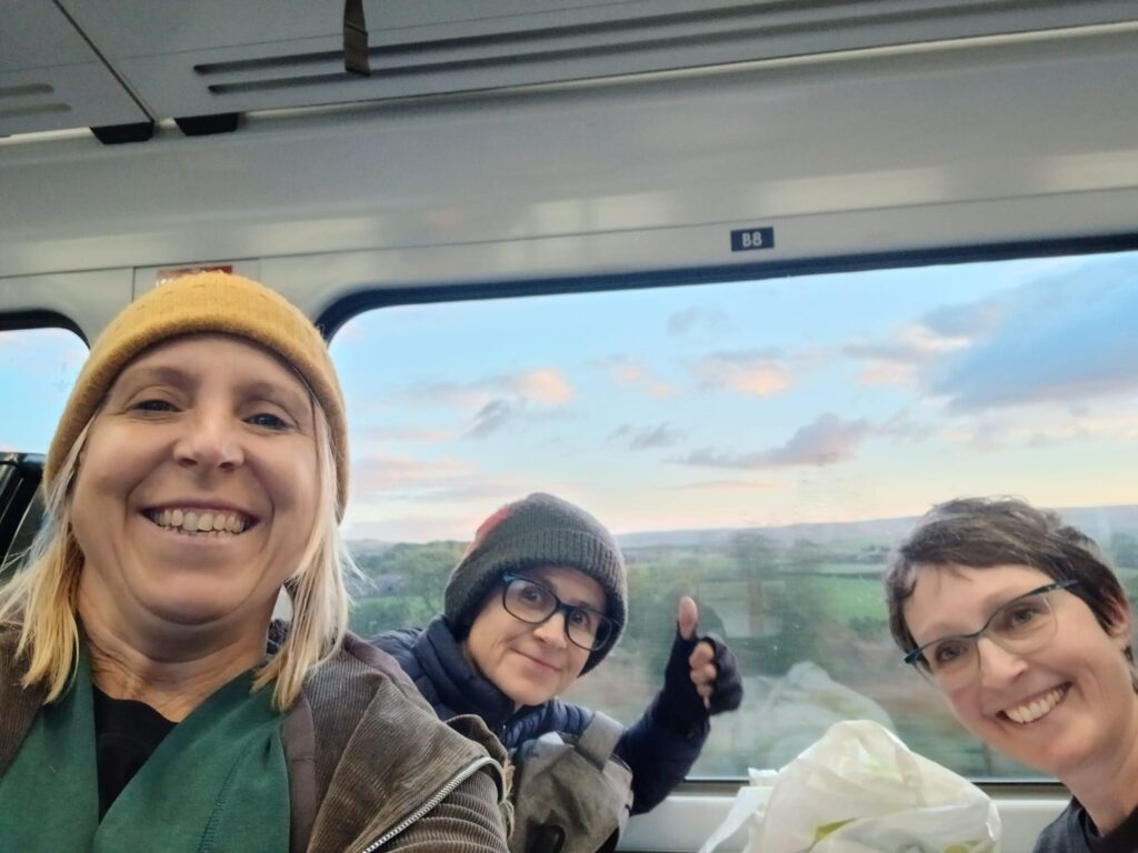 Sarah Hehir, Verity Healey and Becky on a train with countryside through the window. They are smiling and Verity has her thumb up.