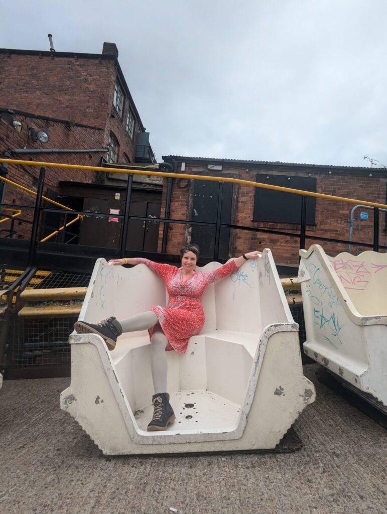 Becky in an old waltzer car outside Beaver Works. Her arms are stretched over the top of the car, one of her feet up on the front. She is wearing a red and grey patterned dress.